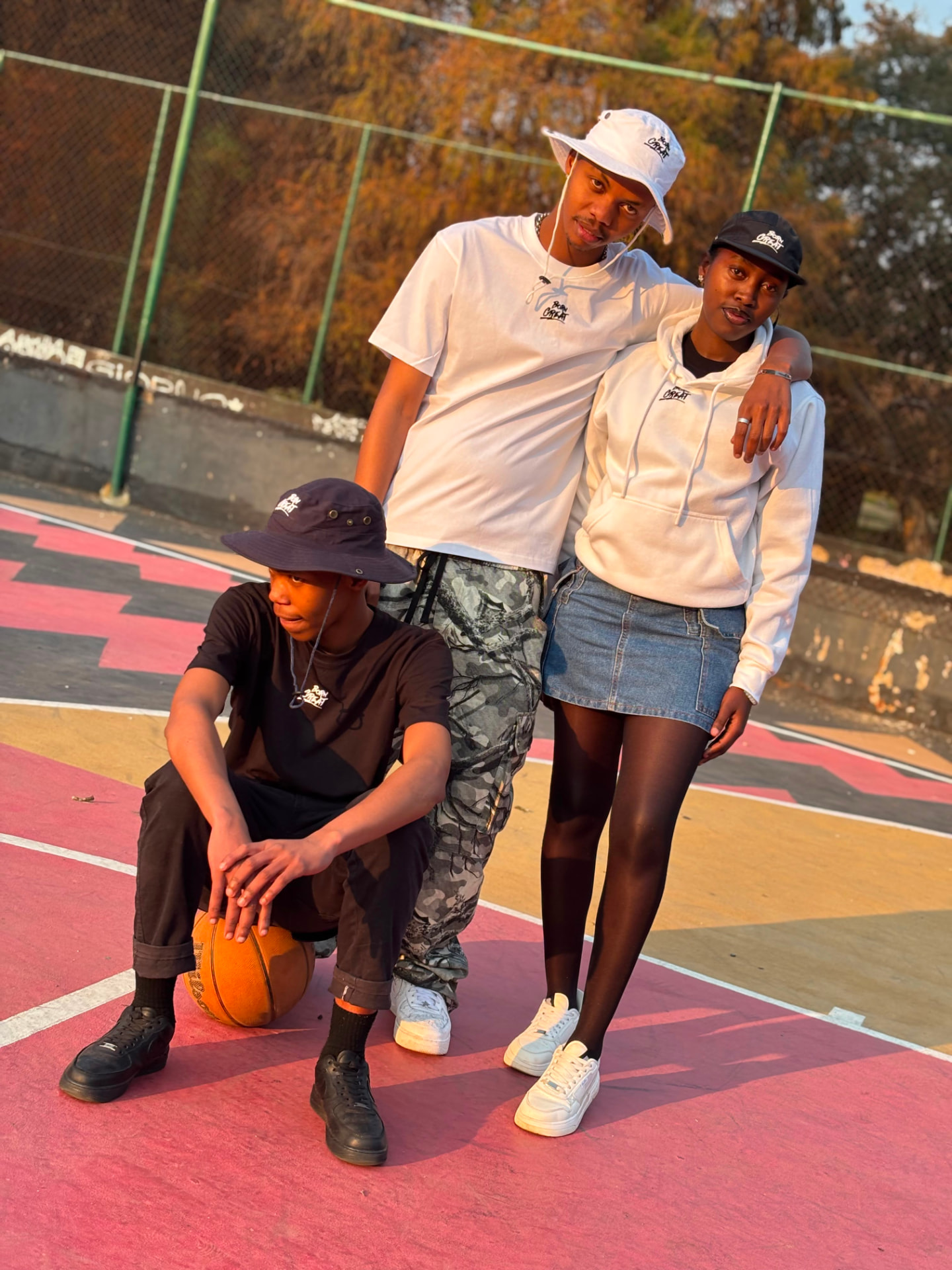 Three friends on a painted basketball court in Born Great white tees, hoodie and bucket hats